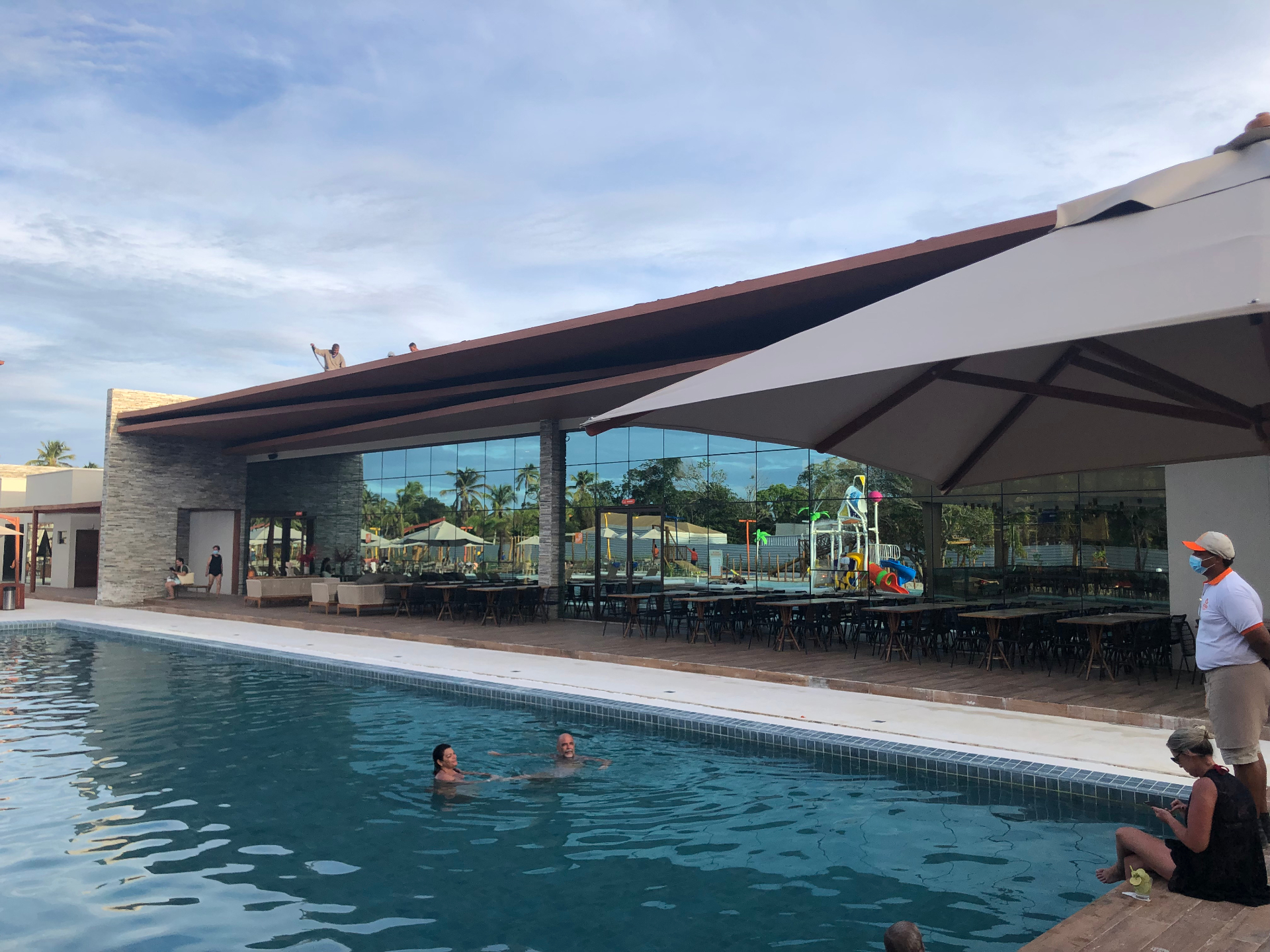 Fachada de clube com grande cobertura, fechamento em vidro e piscina em primeiro plano